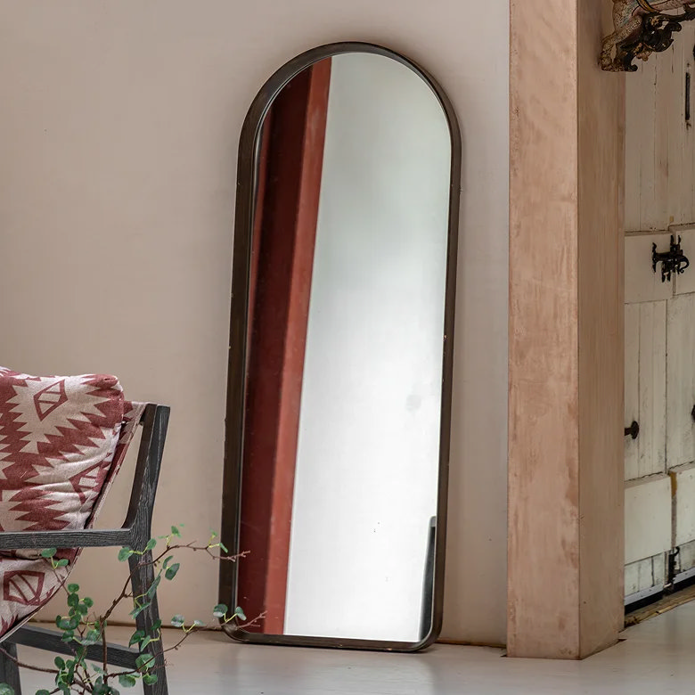 Tall arched floor mirror with dark wood frame leaning against a living room wall, reflecting light and adding height to the space.