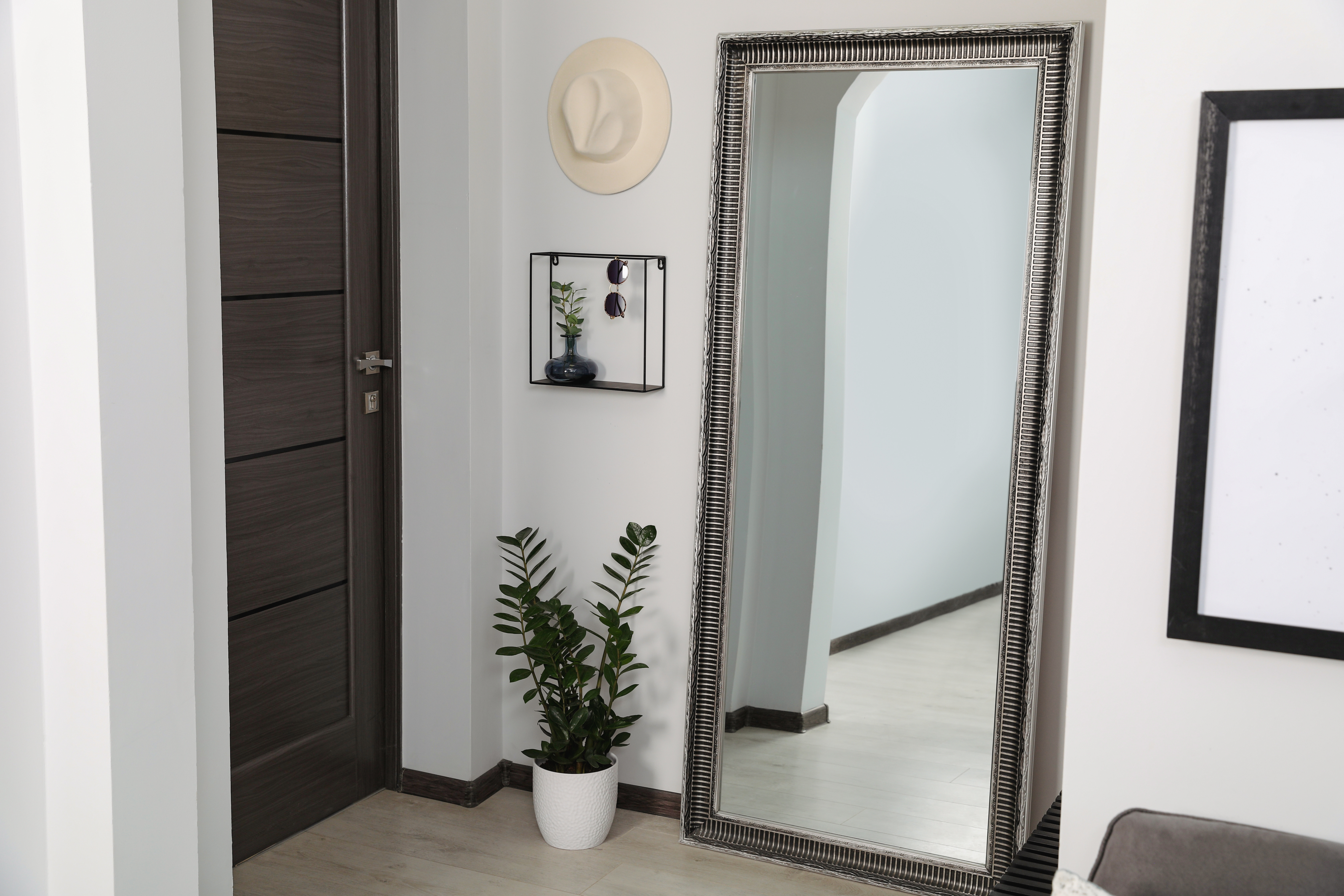 Leaning full-length mirror brightening a small hallway with dark wood door, wall shelf and plant—using mirrors to make a narrow hall look wider.