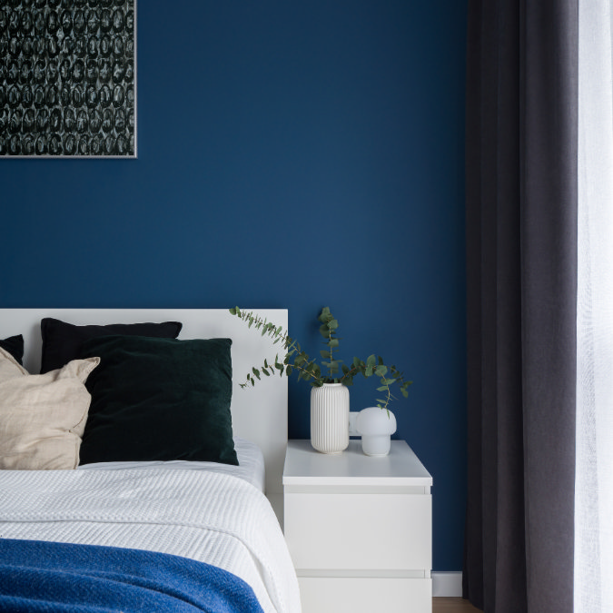 Bedroom with deep blue wall, dark curtains and layered cushions creating a warm, cocooned feel