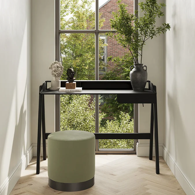 Modern black writing desk with green stool and decorative vase by a tall window with garden view.