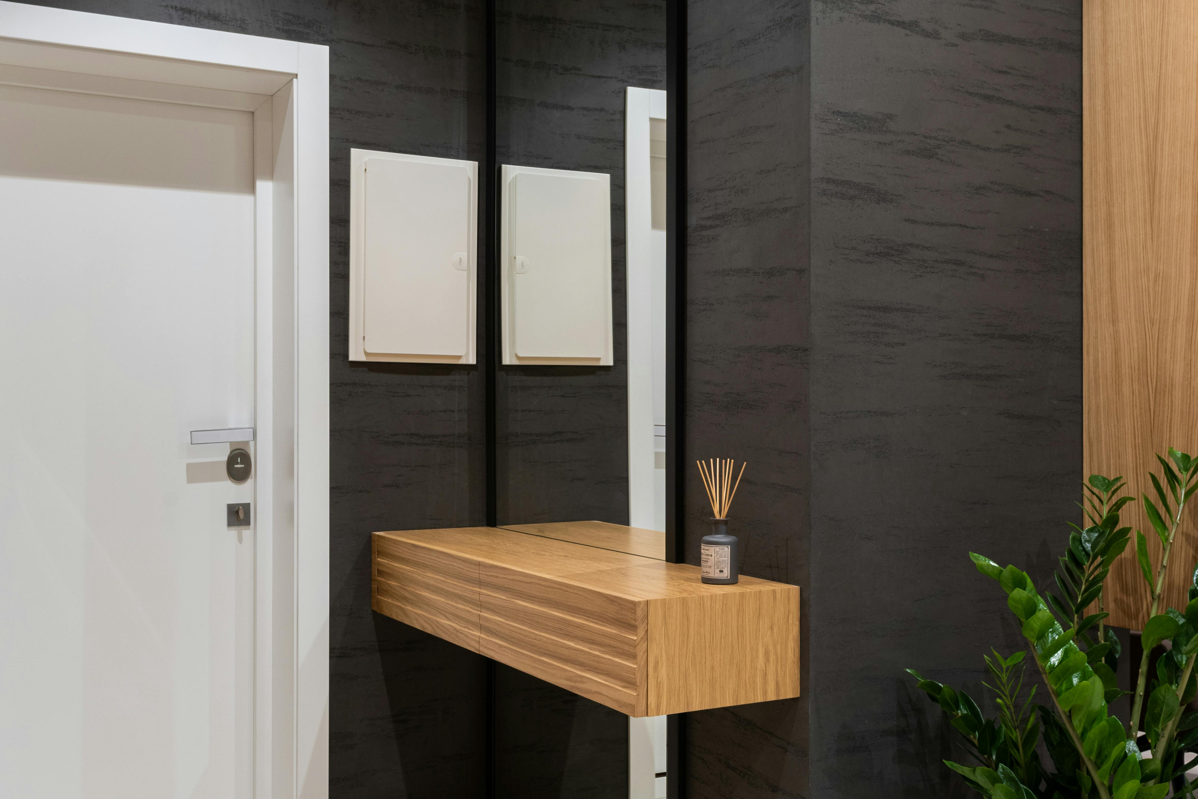 Narrow hallway idea with floor-to-ceiling mirror and floating wood console shelf, reed diffuser and plant—dark walls and white door for a wider, uncluttered look.