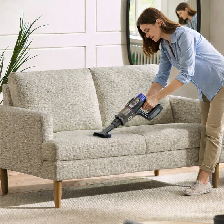 Woman vacuuming a light fabric sofa as part of a living room spring cleaning routine