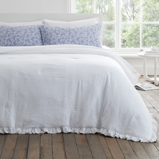 Freshly‐made white linen bed with ruffled duvet and lavender floral pillowcases in a bright, airy bedroom with rustic wooden floor.