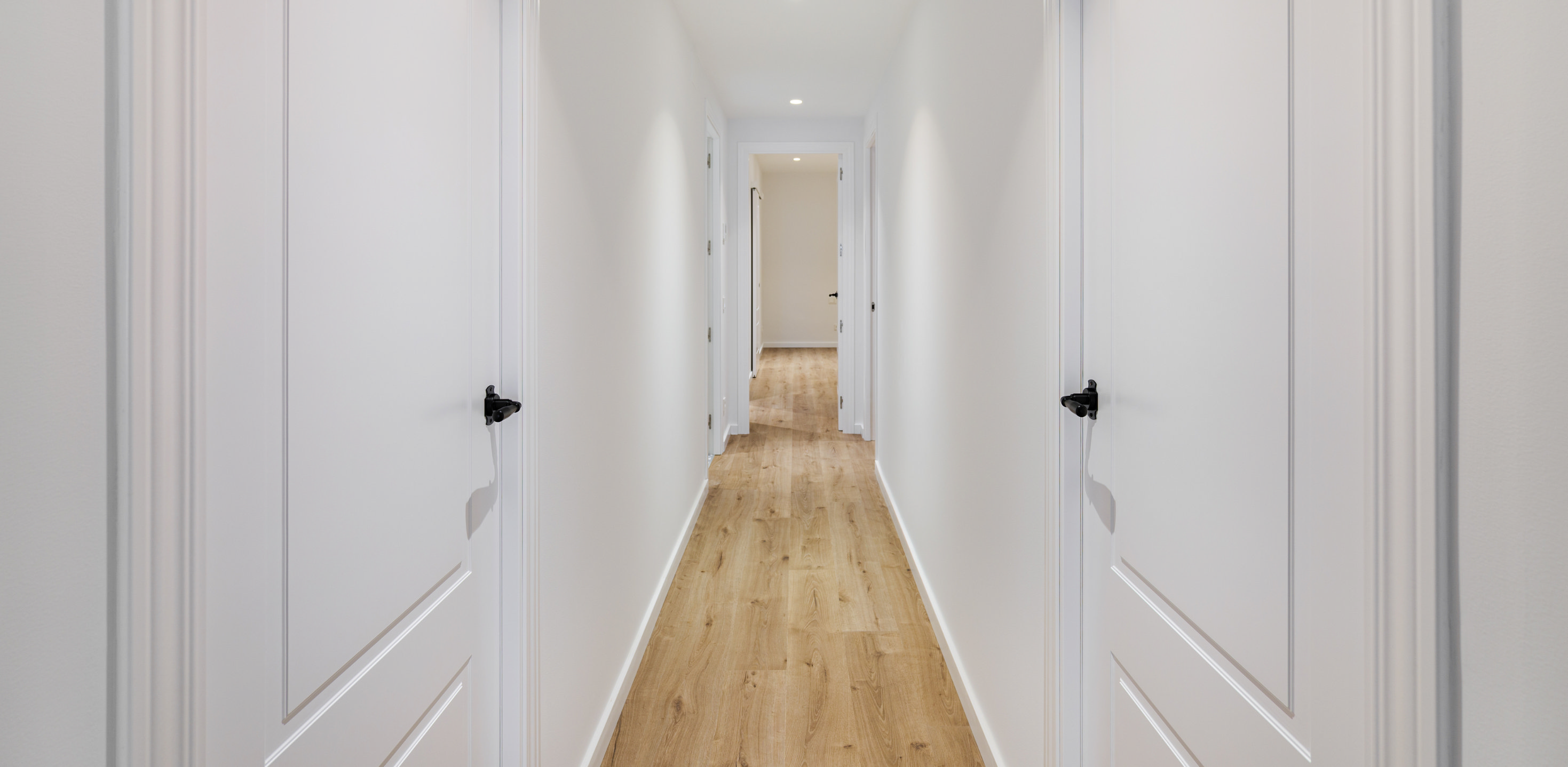 Bright white narrow hallway with light oak floorboards and evenly spaced downlights that visually widen the corridor.  