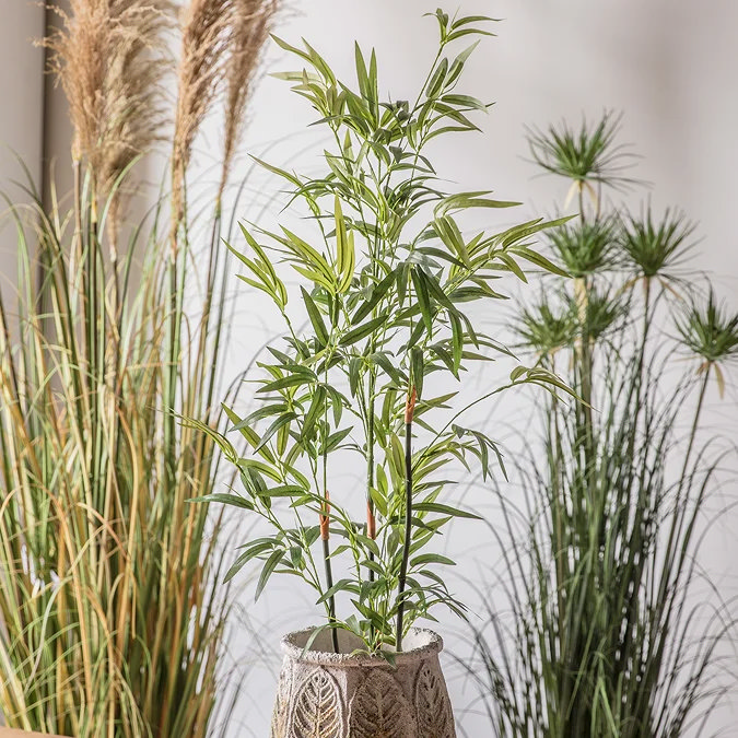 Faux bamboo plant in leaf-patterned stone pot, styled with pampas grass and papyrus for layered indoor greenery backdrop.
