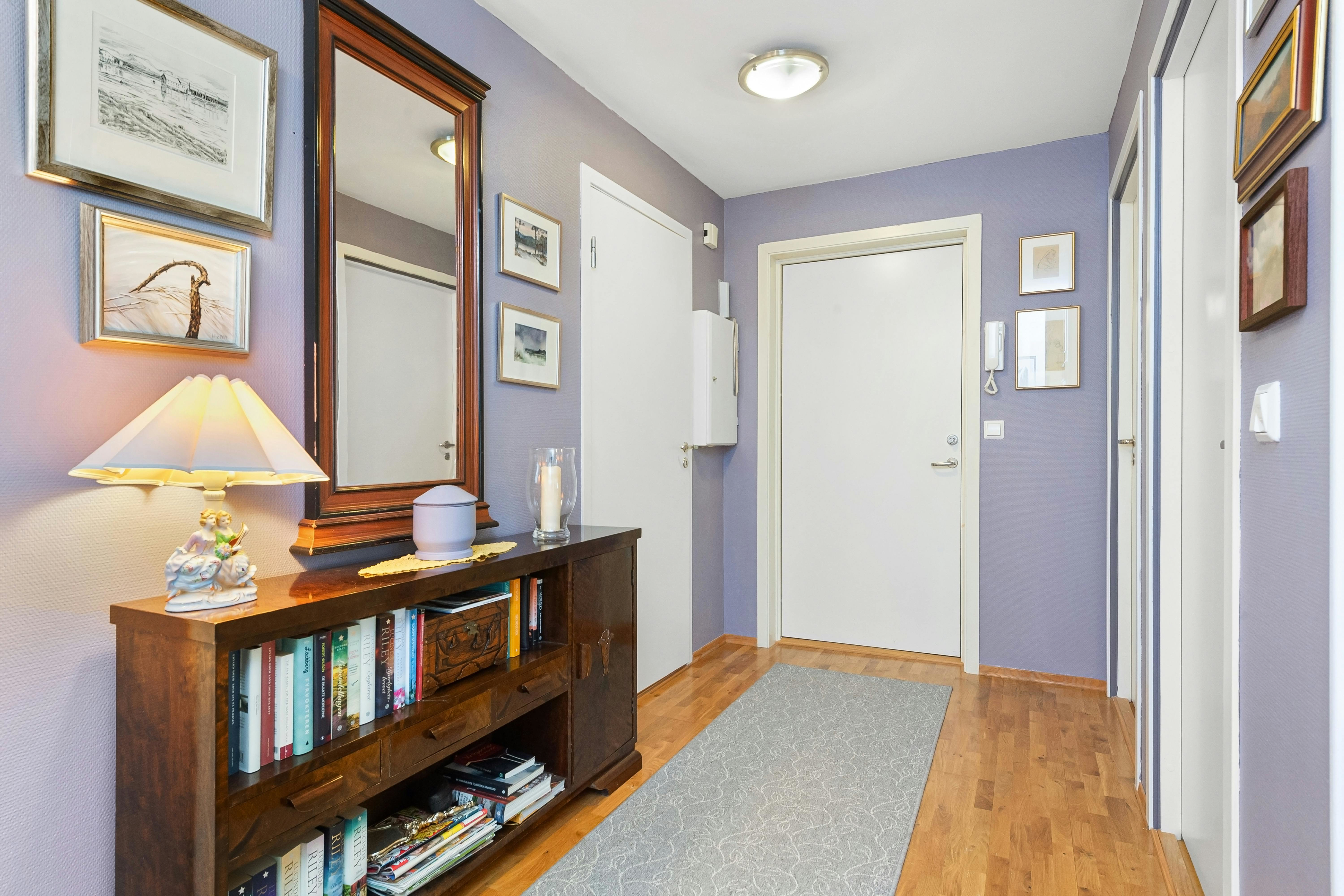 Narrow hallway with lilac walls, slim console and large mirror, gallery art and a long runner on wood flooring—lighting and decor tricks to make halls look wider.