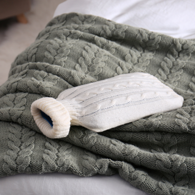 White hot water bottle with knitted cover resting on a chunky green blanket