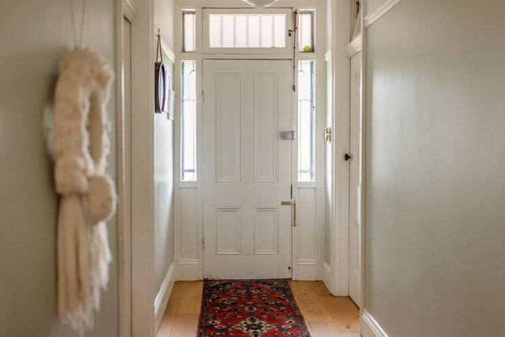 Narrow hallway with white front door, glass sidelights and transom, pale walls and a slim red runner on wood flooring to maximise light and width.   