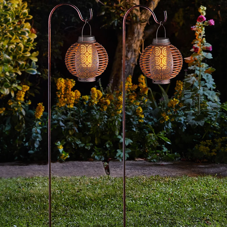 Decorative garden lanterns hanging from shepherd’s hook stakes beside a flower border at night