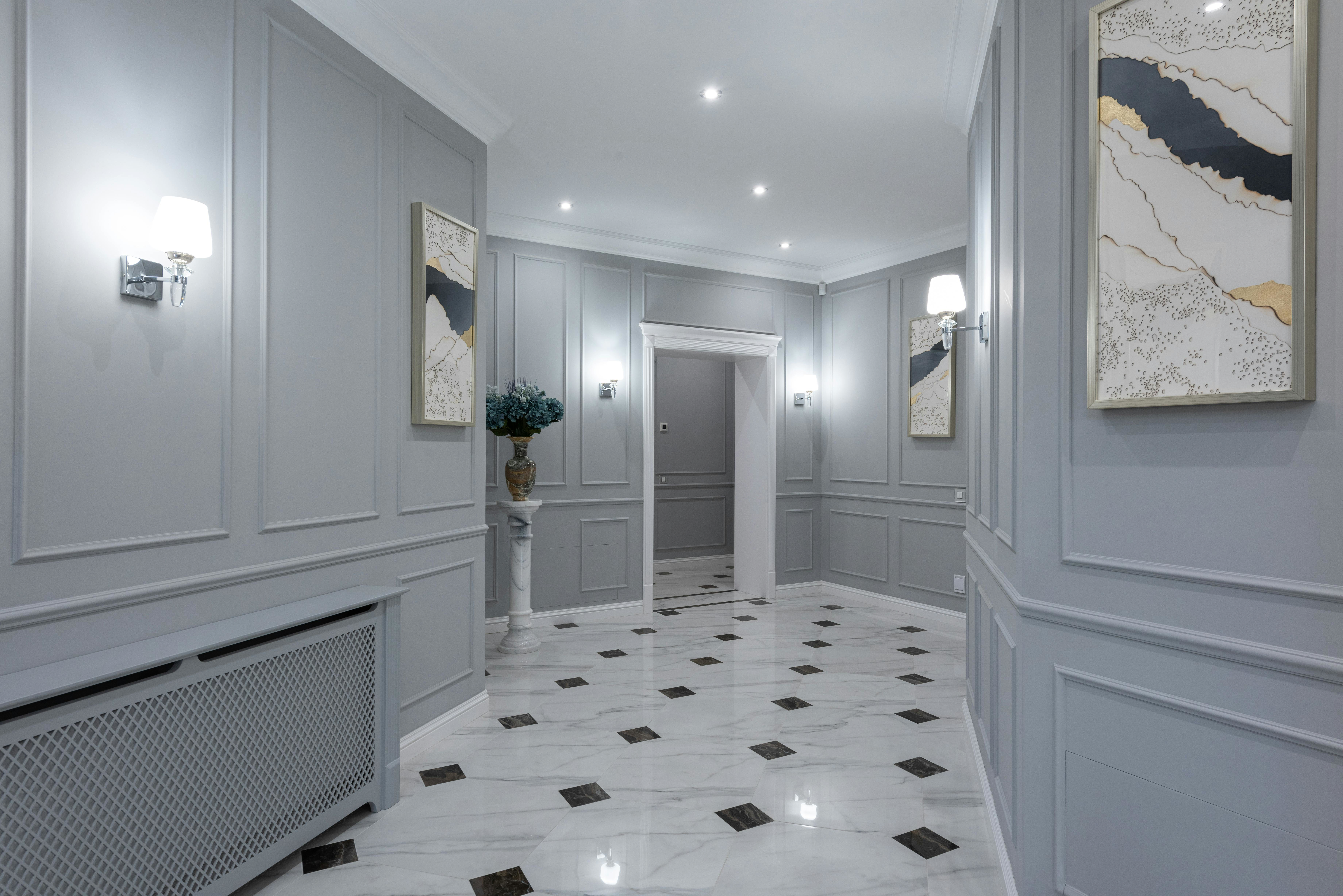 Grey panelled hallway with wall sconces and glossy marble floor, layered lighting that reflects and makes a narrow space feel wider.