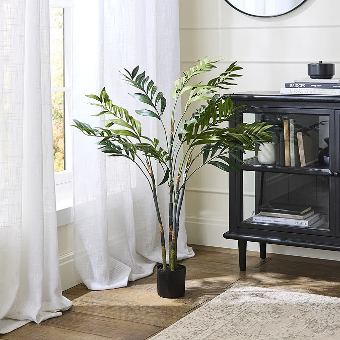 Tall faux areca palm in a black pot adding spa-like greenery beside a window and glass-front cabinet.