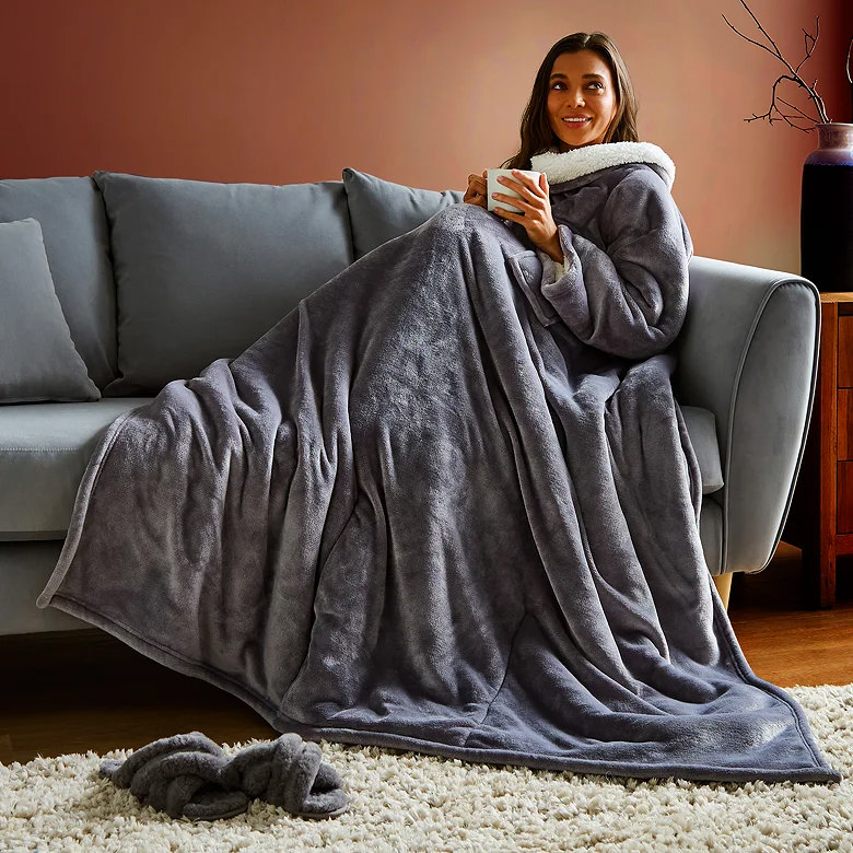 Woman relaxing on sofa wrapped in grey wearable blanket holding mug