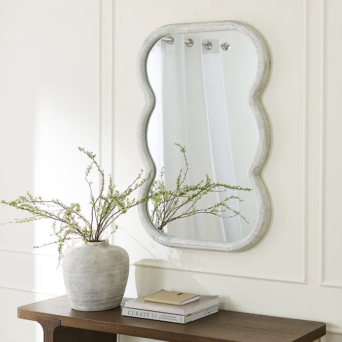 Ripple‑edge, concrete‑look wall mirror reflecting white curtains, styled above a dark wood console with stone vase and greenery – ideal industrial bedroom accent.
