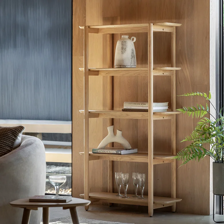 Light oak ladder-style shelving unit with books, ceramics and glassware in a modern living room.