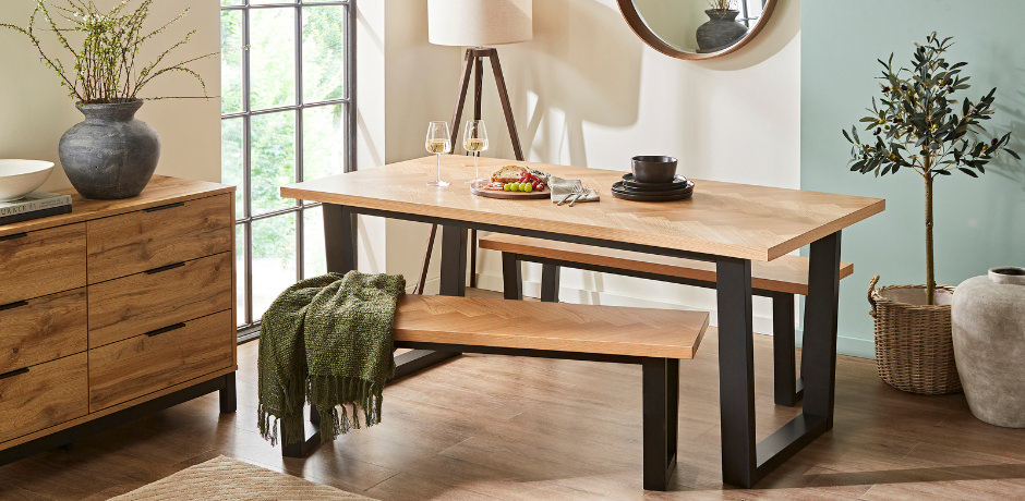 Modern dining room with herringbone oak dining table and matching bench, featuring black metal legs, set beside a sideboard, indoor plants, and soft natural lighting from a large window.