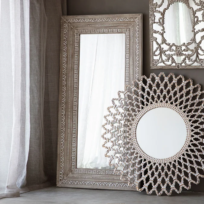 Tall rectangular floor mirror with ornate carved wooden frame in distressed finish, leaning against a living room wall to add light and elegance.