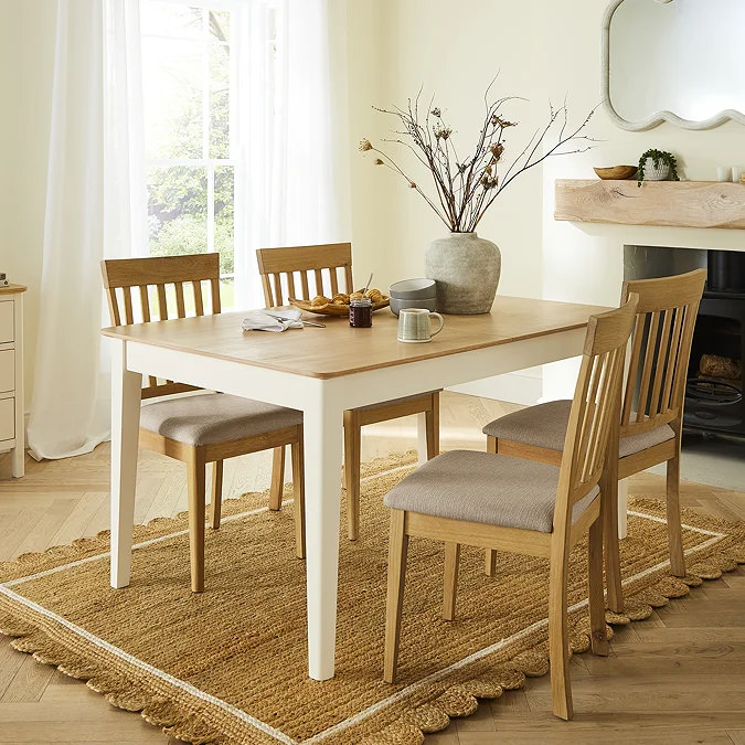 Bright and airy dining room with a two-tone wooden table, four slatted back dining chairs, and a textured jute rug beneath, styled with ceramic mugs and a rustic stone vase of dried branches.