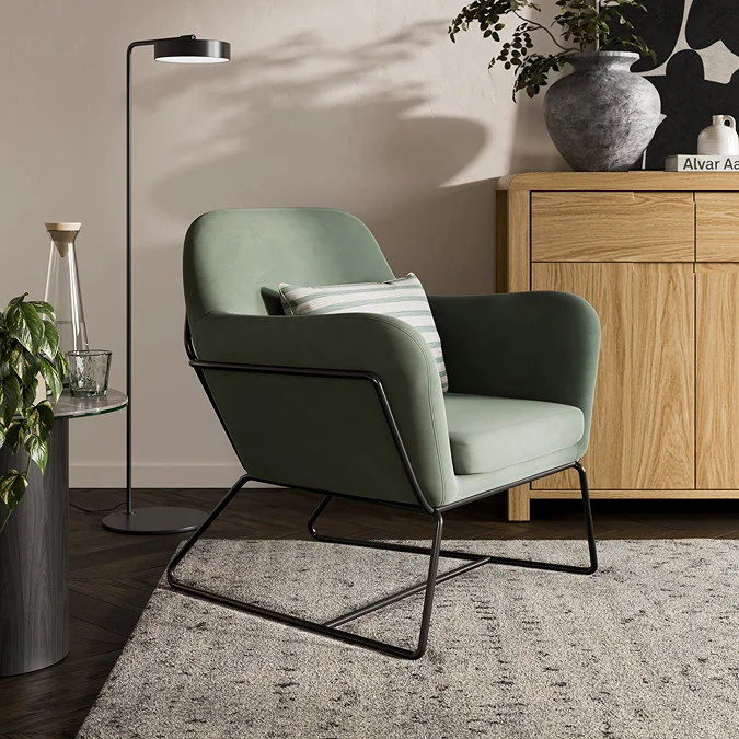 Sage green accent chair with black metal frame sitting on a soft grey patterned rug beside a wooden sideboard and floor lamp, showing a calm, modern home office reading corner setup
