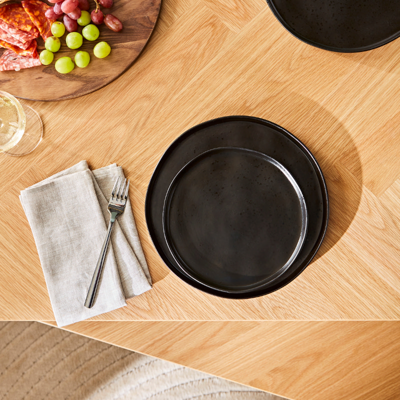 Close-up of a modern dining setup with matte black stoneware plates, neutral linen napkin, and stainless steel fork on a herringbone oak dining table, with charcuterie board and wine glass nearby.