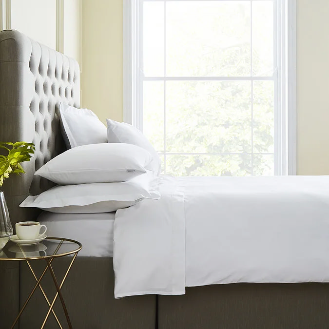 Crisp white hotel-style sheets and neatly stacked pillows on a taupe tufted bed next to a slim brass bedside table.