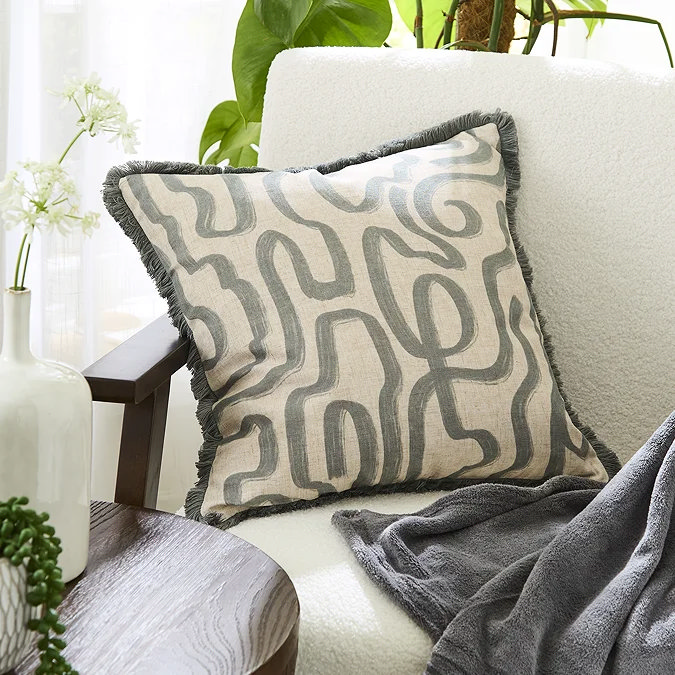Square linen-look cushion with grey abstract maze pattern and fringe piping on a cream bouclé armchair beside a round wooden side table.