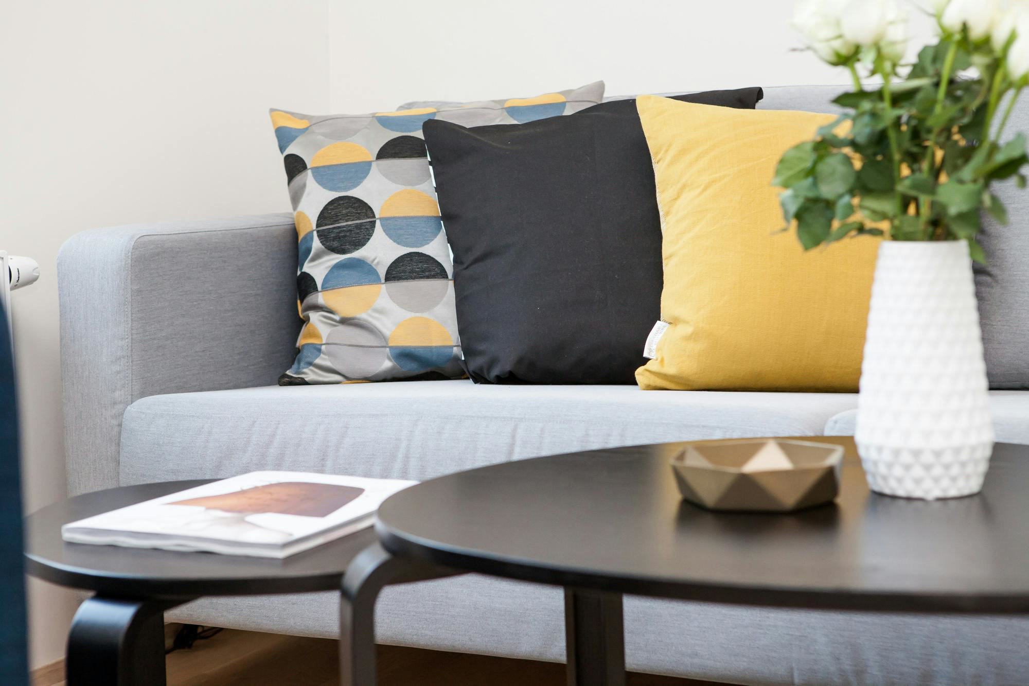 Grey two‑seater sofa styled with three cushions—one geometric grey/blue/yellow, one black, one mustard behind black nesting tables with white rose vase
