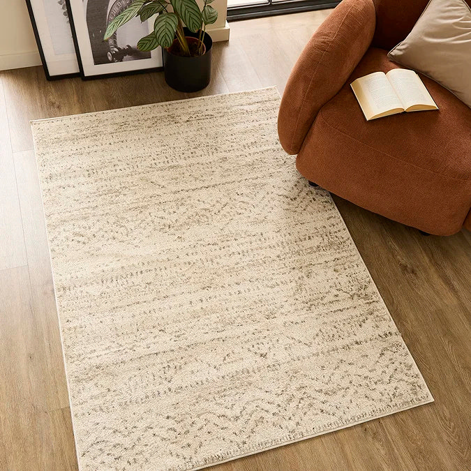 Warm oatmeal-toned patterned rug on wood flooring beside a rust armchair and indoor plant — a light neutral office rug idea that brightens a small workspace, defines a cosy reading or work zone, and keeps the look calm and inviting without visual clutter