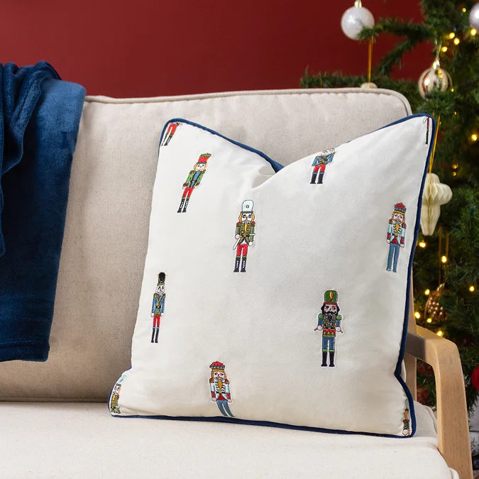 White Christmas cushion with embroidered nutcracker design on beige sofa beside Christmas tree