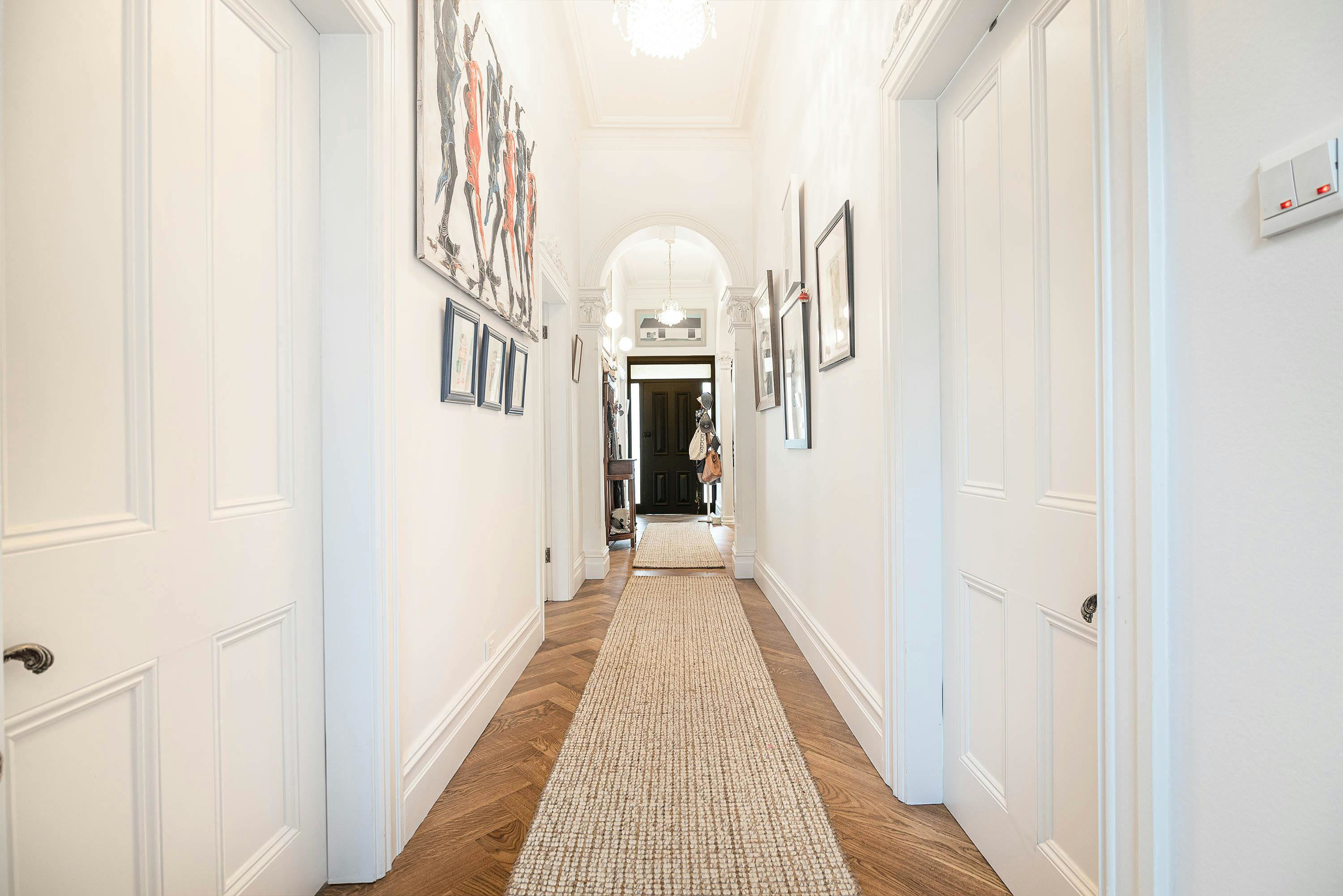 Bright narrow hallway with white walls, gallery art, wall lights and a long runner on herringbone wood flooring leading to an arched doorway—styling that makes a corridor feel wider. 