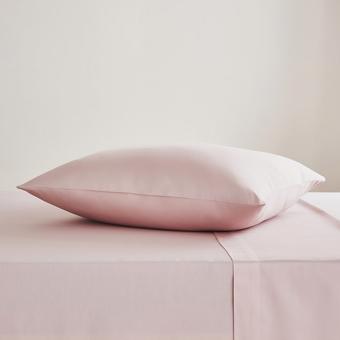 square coloured pillow in pale pink pillowcase, resting on matching flat sheet on bed