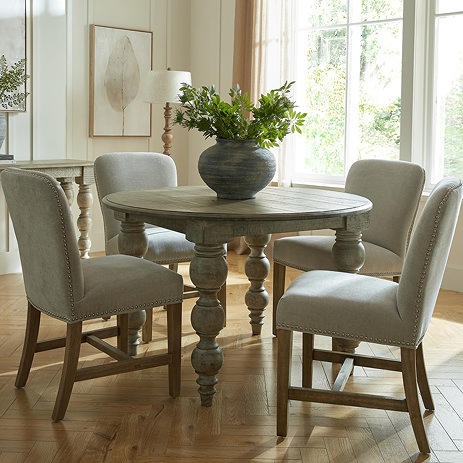 image of rustic dark brown dining table in round shape, with carved legs and grey upholstered dining chairs
