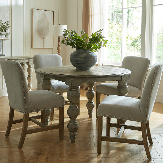 image of rustic dark brown dining table in round shape, with carved legs and grey upholstered dining chairs