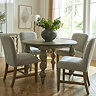 image of rustic dark brown dining table in round shape, with carved legs and grey upholstered dining chairs