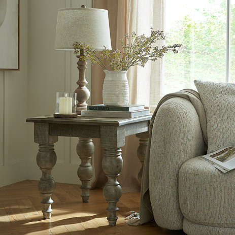 image of square rustic side table with carved legs, topped with matching lamp, next to grey upholstered sofa