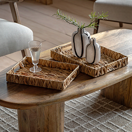 pair of shallow woven rectangle trays on an oval coffee table, with glass and faux plants displayed in them