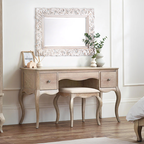 ornate french inspired dressing table pushed up against white wall with intricate mirror above, complete with matching light wood fabric stool
