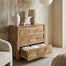 side on image of three drawer chest in natural colour with arch design on the front, bottom drawer pulled out to show clothes stored within