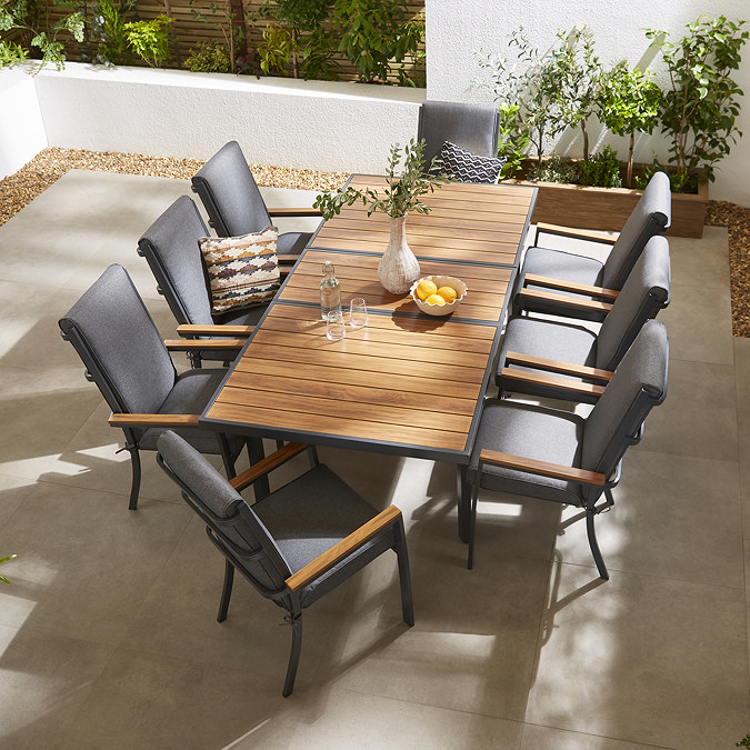 image taken o a patio from above of teak finish slatted outdoor dining table, with eight matching grey chairs with teak arms tucked around it