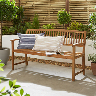 image of three seater bench in oil teak finish, with patterned cushions, in front of a white wall in a garden