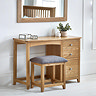 image of solid oak single dressing table with grey padded stool tucked underneath, pushed up against light grey wall with wooden mirror hanging above