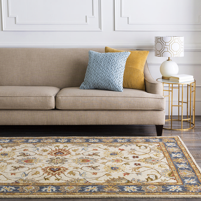 image of vintage tan coloured rug with brown, dark red, yellow and blue floral patterns, on floor in front of brown sofa