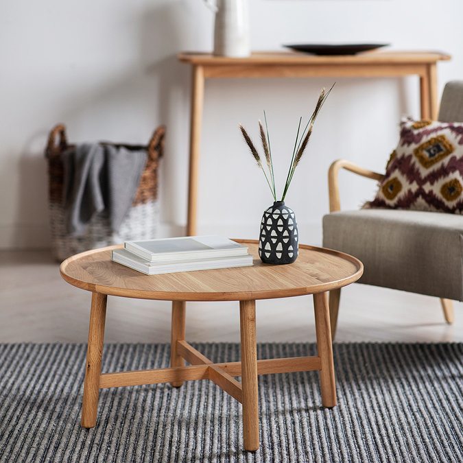 image of round oak coffee table with two books and a faux plant, on top of a black and white striped rug next to a light grey armchair