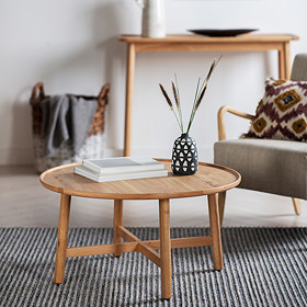 image of round oak coffee table with two books and a faux plant, on top of a black and white striped rug next to a light grey armchair