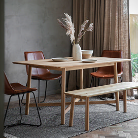 solid oak wooden dining table with matching bench, dressed up with plates and faux plant, on a grey rug next to a big window