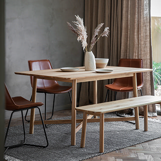 solid oak wooden dining table with matching bench, dressed up with plates and faux plant, on a grey rug next to a big window