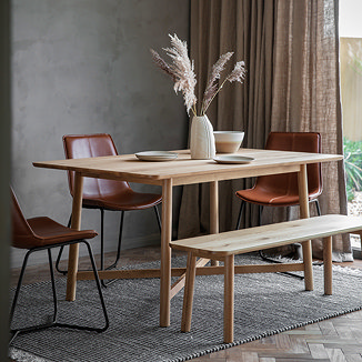solid oak wooden dining table with matching bench, dressed up with plates and faux plant, on a grey rug next to a big window