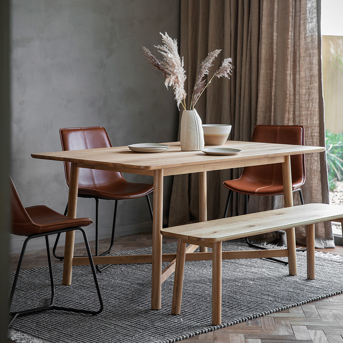 solid oak wooden dining table with matching bench, dressed up with plates and faux plant, on a grey rug next to a big window
