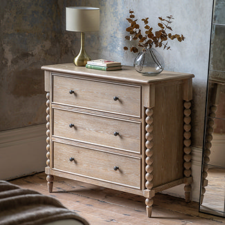image of limed oak three drawer chest with hand-crafted bobbin detail, against a distressed grey wall and topped with a lamp and faux plant