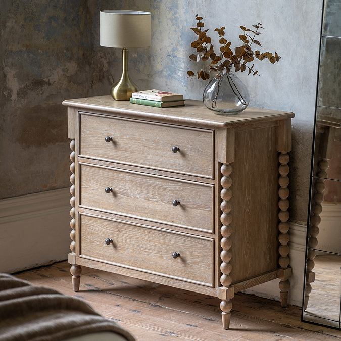 image of limed oak three drawer chest with hand-crafted bobbin detail, against a distressed grey wall and topped with a lamp and faux plant