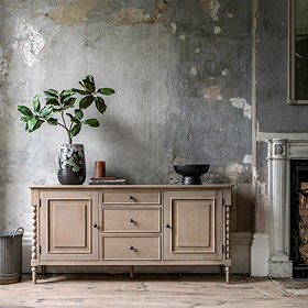 image of limed oak sideboard with two doors on either side and three drawers in the middle, with faux plants on top, pushed against a distressed grey-blue wall